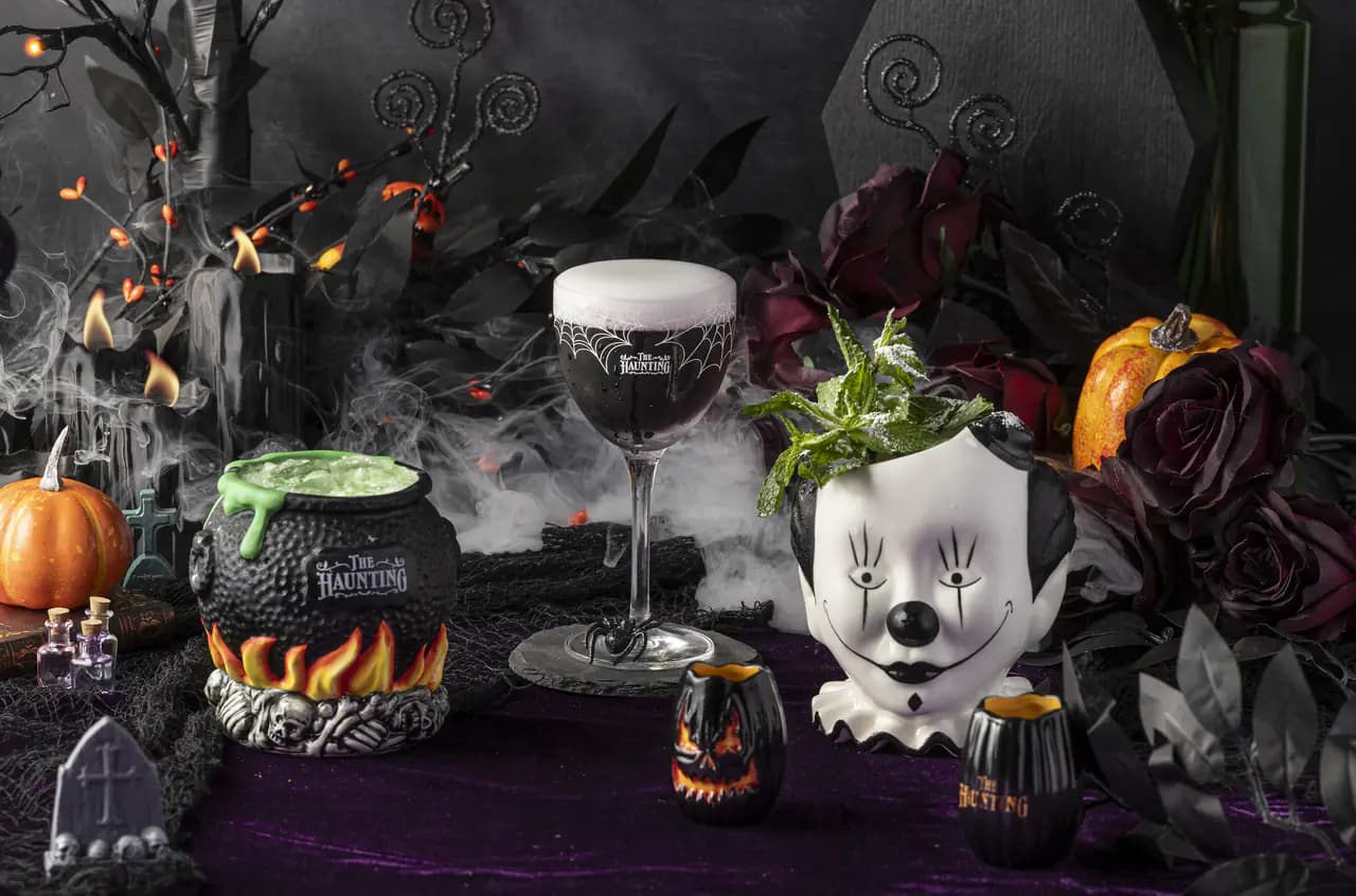 Halloween cocktails in a cauldron mug, a stemmed glass with a spiderweb design and a clown-head mug, surrounded by pumpkins, dark roses and fog