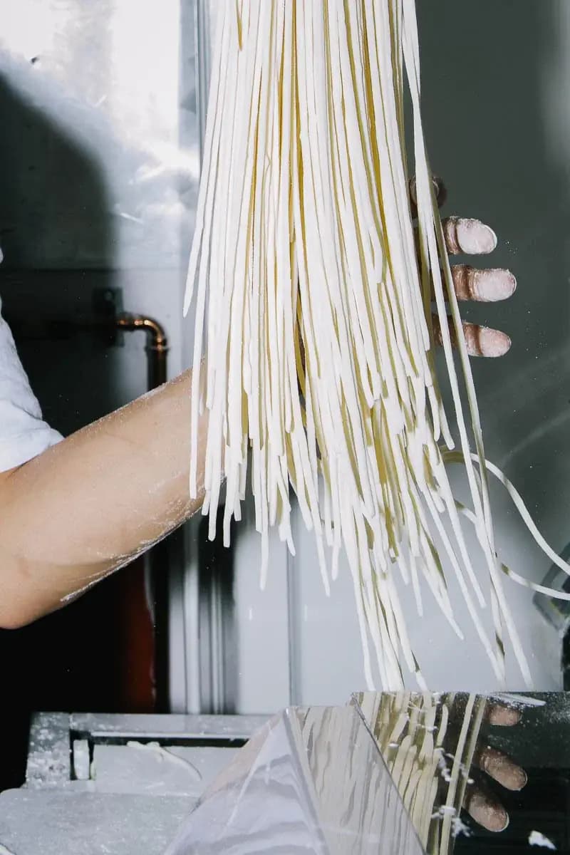 A cook lifts a bundle of freshly cut, flour-dusted udon noodles from a noodle machine
