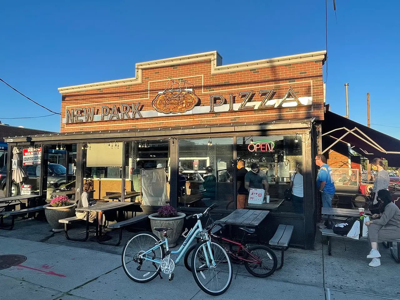 The brick storefront of New Park Pizza on Cross Bay Boulevard in Howard Beach, with its sign, outdoor picnic tables and bicycles parked in front