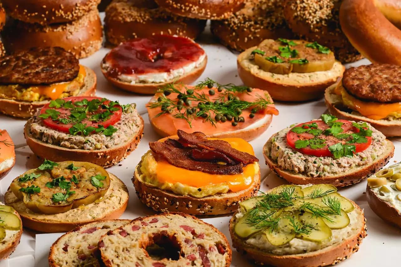 An overhead spread of open-faced bagel sandwiches topped with smoked salmon, bacon, pickled cucumbers, tomatoes and fresh dill, with whole sesame and everything bagels at the edges