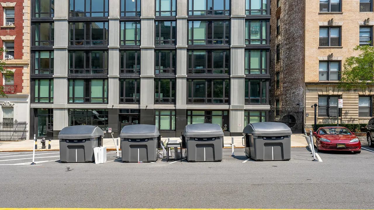 Four large gray Empire Bins trash containers lined up in the curb lane in front of apartment buildings