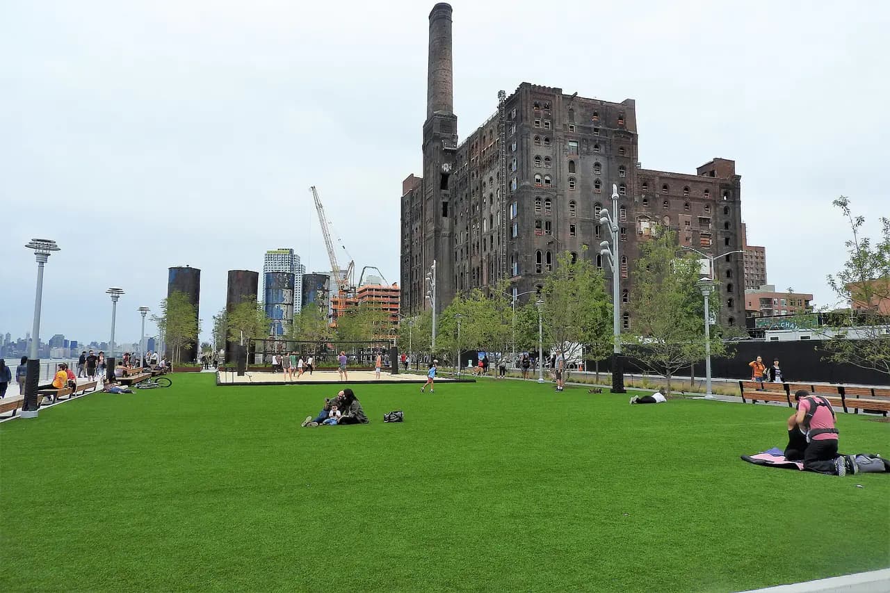 A wide lawn at Domino Park in Williamsburg with the old Domino Sugar refinery building and its smokestack behind it