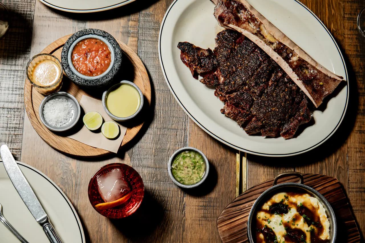 A sliced charred rib eye with a split marrow bone on a white platter, surrounded by salsas, lime, salt, a cocktail and a dish of mashed potatoes
