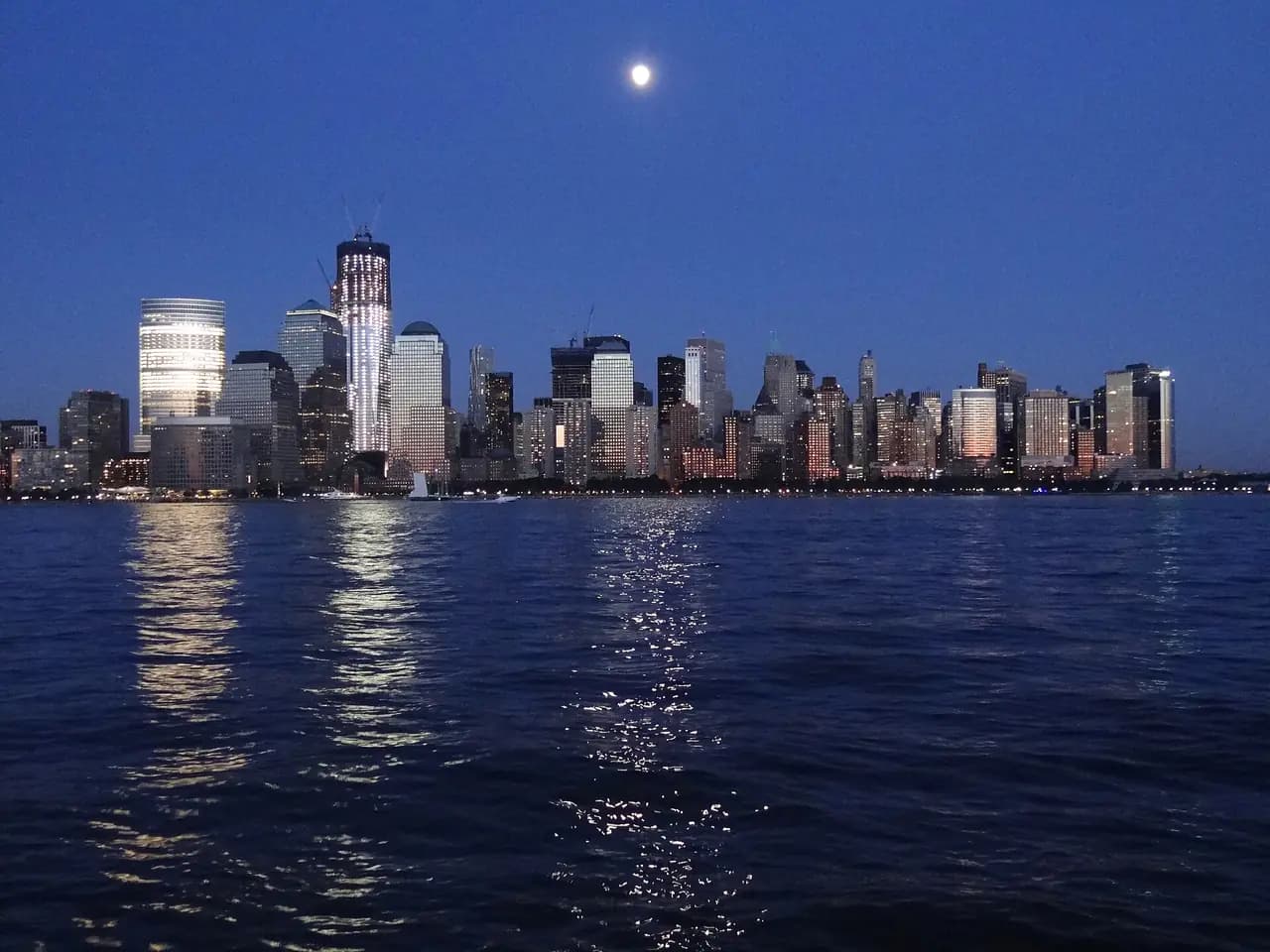 A bright moon in a deep blue evening sky above the lit towers of Lower Manhattan, reflected in the Hudson River