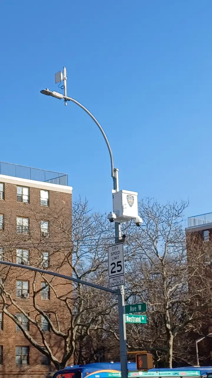 A city speed camera mounted on a pole at the intersection of Avenue W and Nostrand Avenue in Brooklyn, with apartment buildings and bare trees