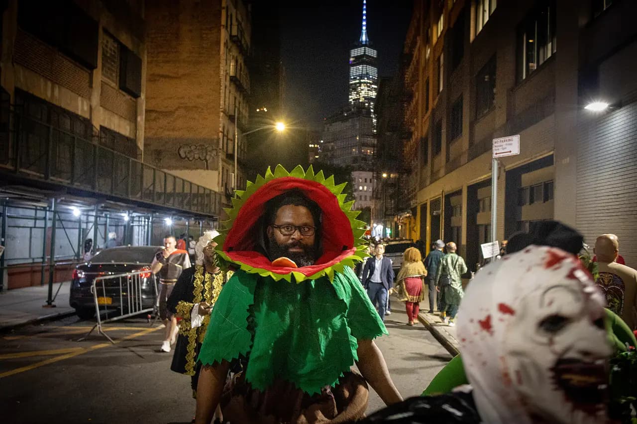 A man in a green Audrey II plant costume walks down a lower Manhattan street at night toward the Village Halloween Parade, with One World Trade Center lit up behind him