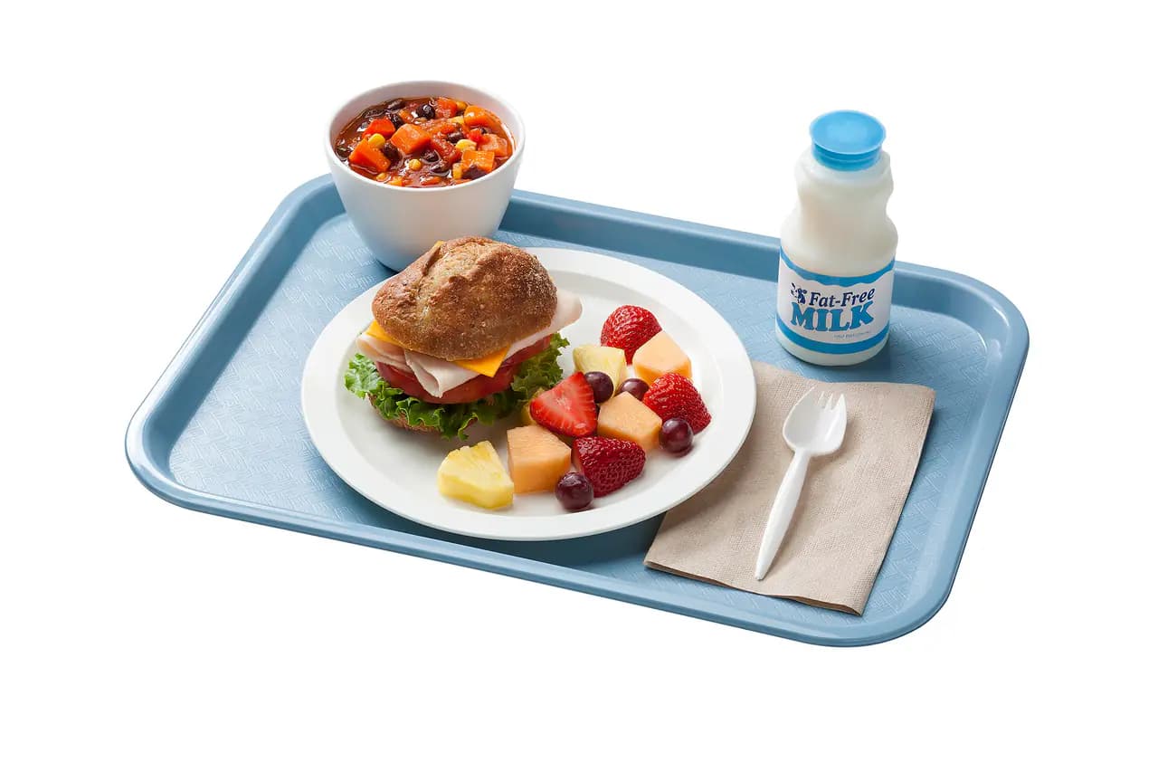 A blue school lunch tray with a sandwich on a whole-grain roll, a cup of bean and vegetable salad, cut fruit and a bottle of fat-free milk