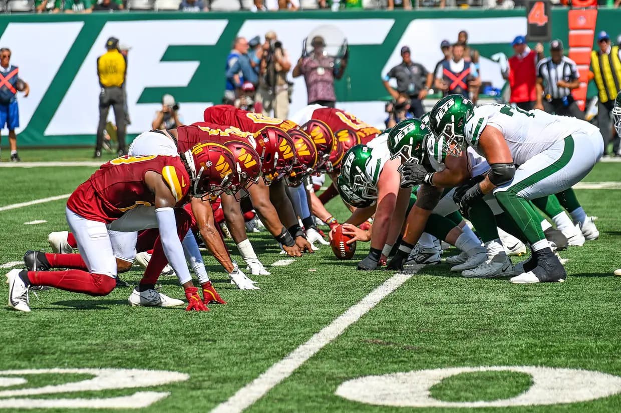 Jets players in white jerseys and Commanders players in burgundy lined up at the line of scrimmage on the field at MetLife Stadium