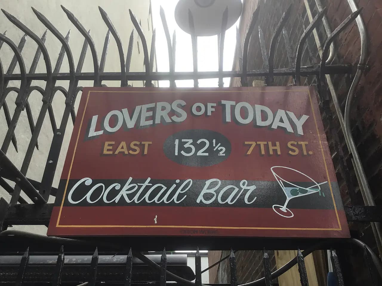 A painted sign reading Lovers of Today, East 132 1/2 7th St., Cocktail Bar, hanging behind an iron fence