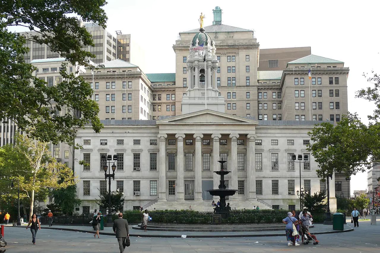 The Greek Revival front of Brooklyn Borough Hall with its columned portico and cupola, a fountain and people walking on the plaza