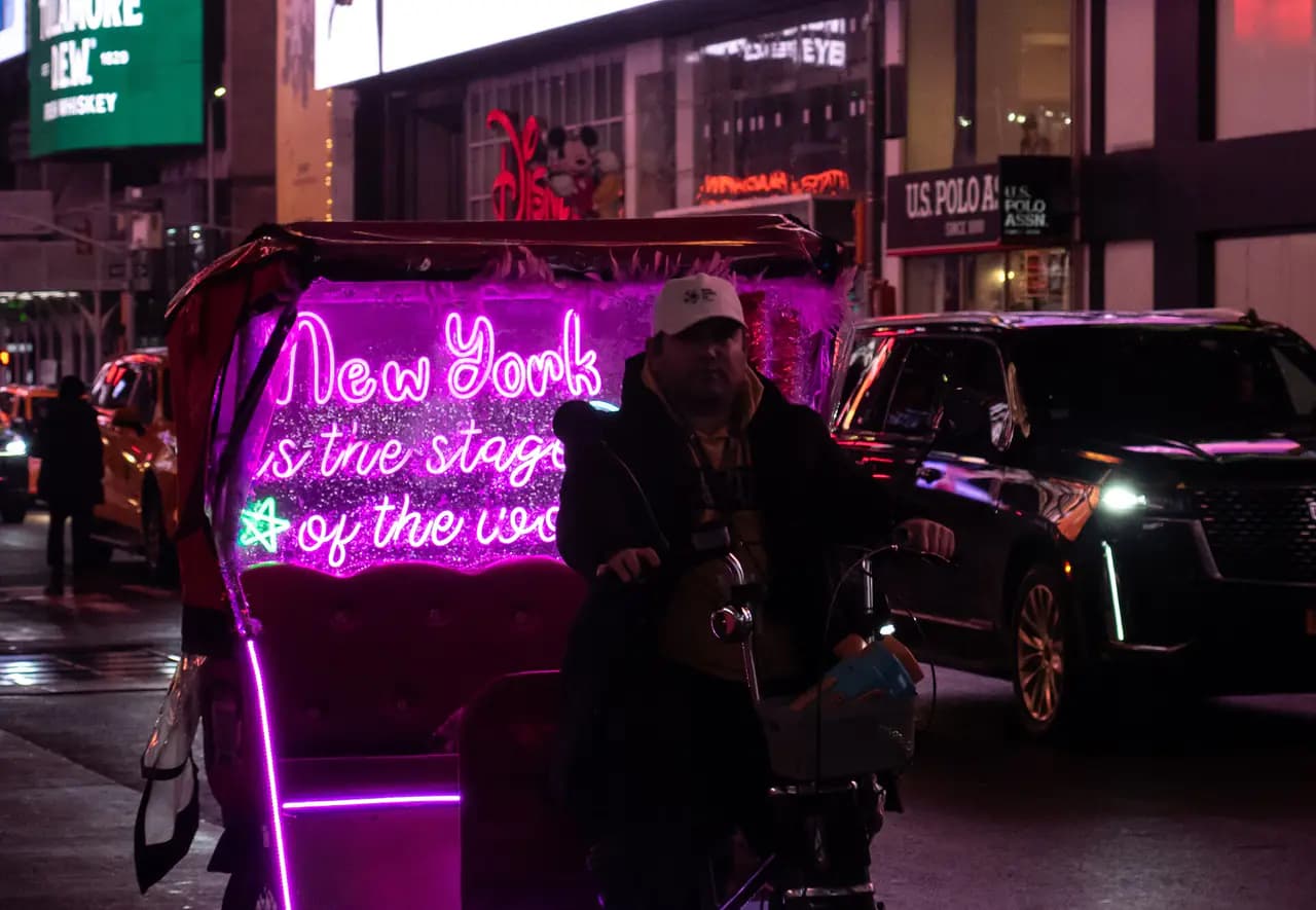 A pedicab lit with pink neon lettering reading 'New York is the stage of the world' waits on a Times Square street at night