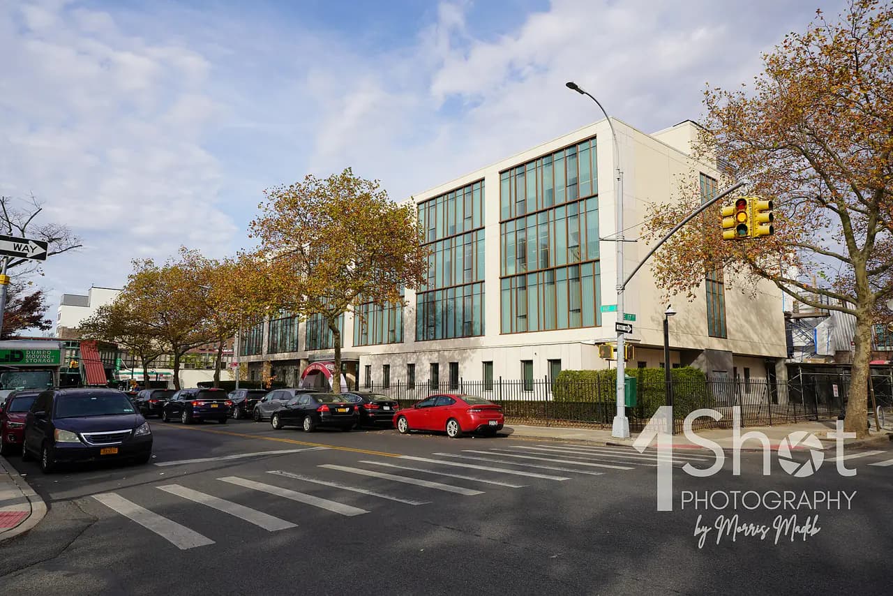 The Yeshivah of Flatbush Joel Braverman High School building on Avenue J in Brooklyn, a modern facade with glass panels, seen from a crosswalk with autumn trees