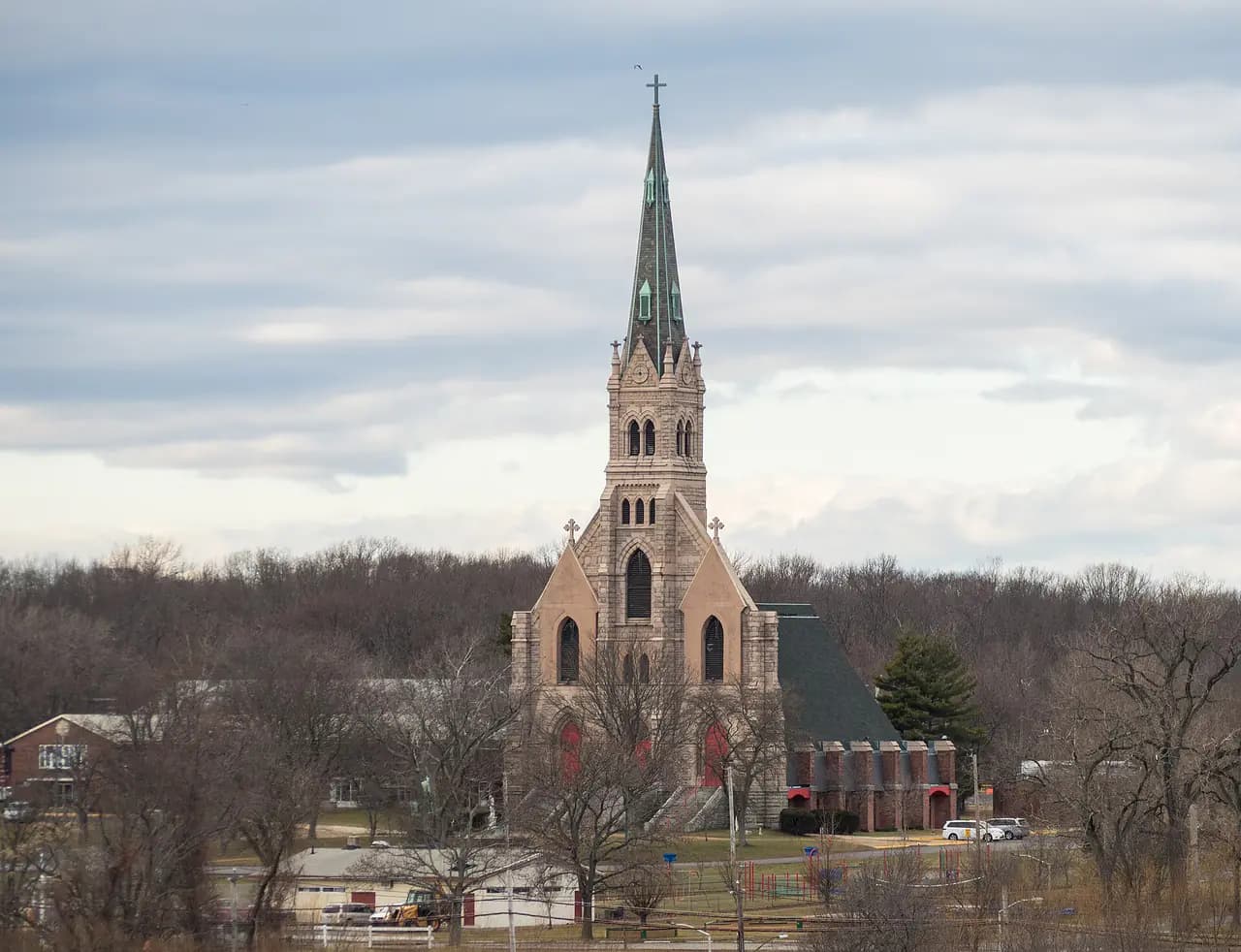 The stone Old Church of St. Joachim and St. Anne with its tall green-tipped spire rising above bare winter trees and low campus buildings at Mount Loretto