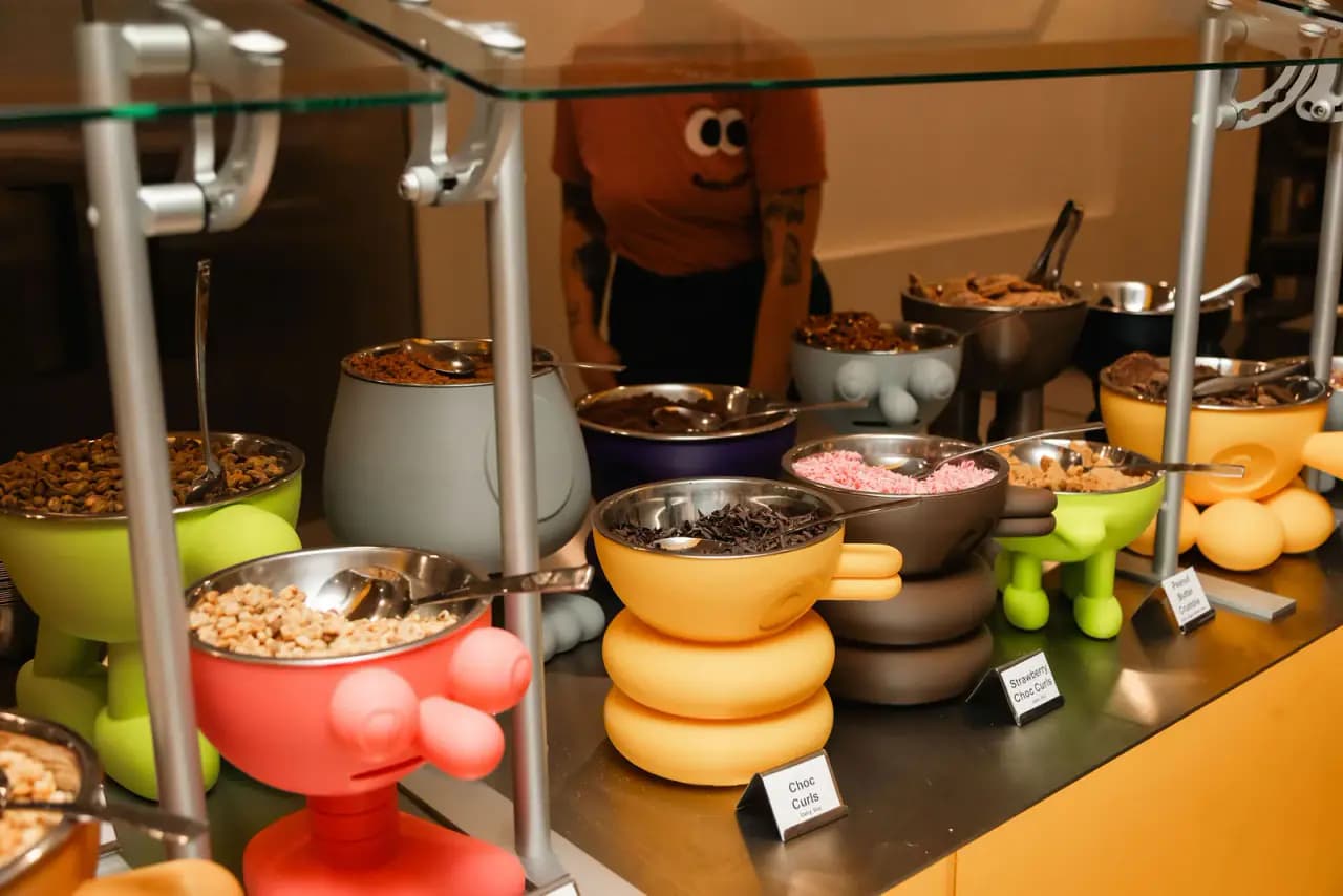 A toppings counter at Softville with colorful character-shaped bowls of sprinkles, chocolate curls and crumbs behind a glass sneeze guard