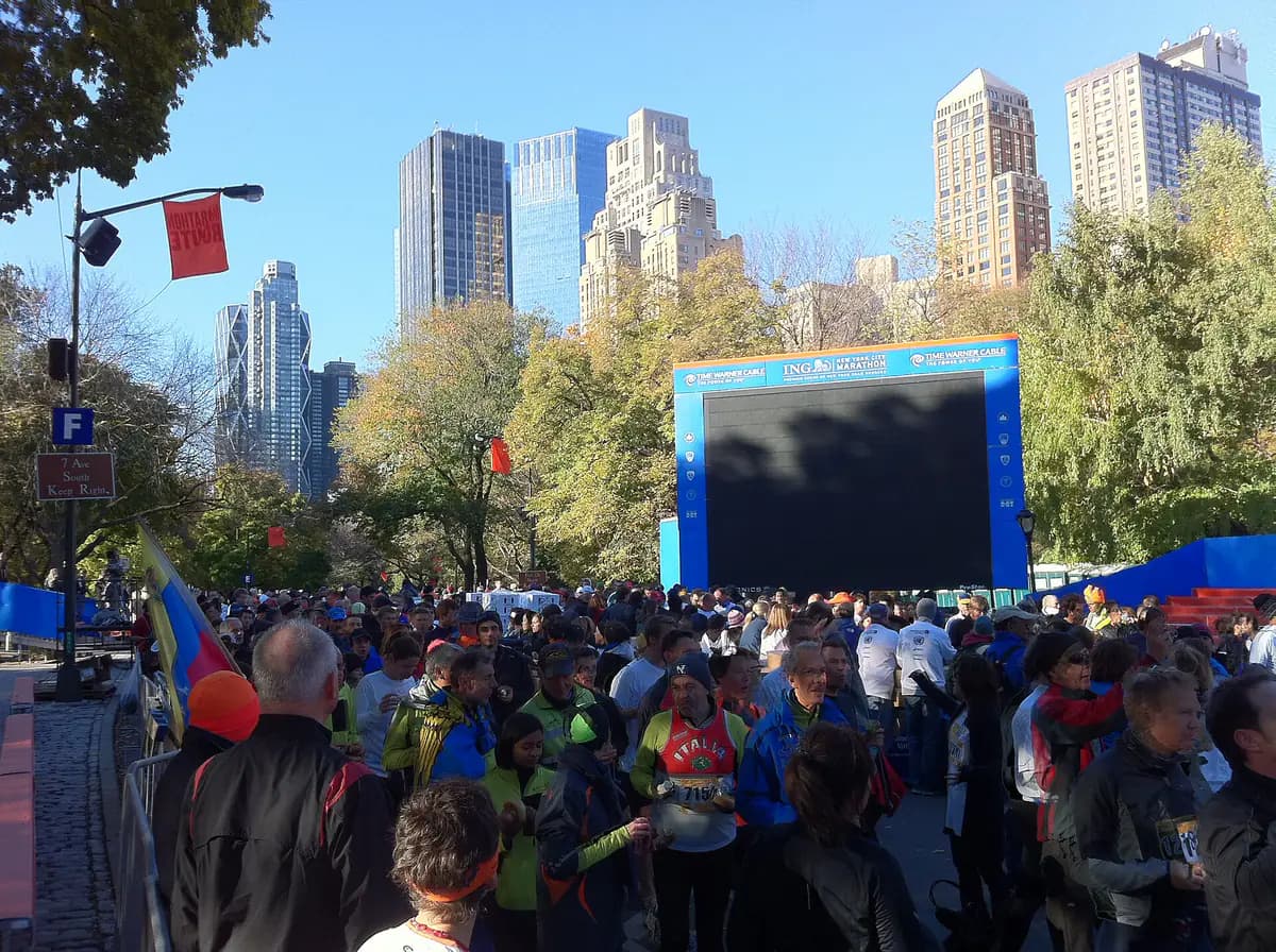 Runners and spectators crowd the finish area of the New York City Marathon in Central Park, with a giant screen and Midtown towers behind the autumn trees