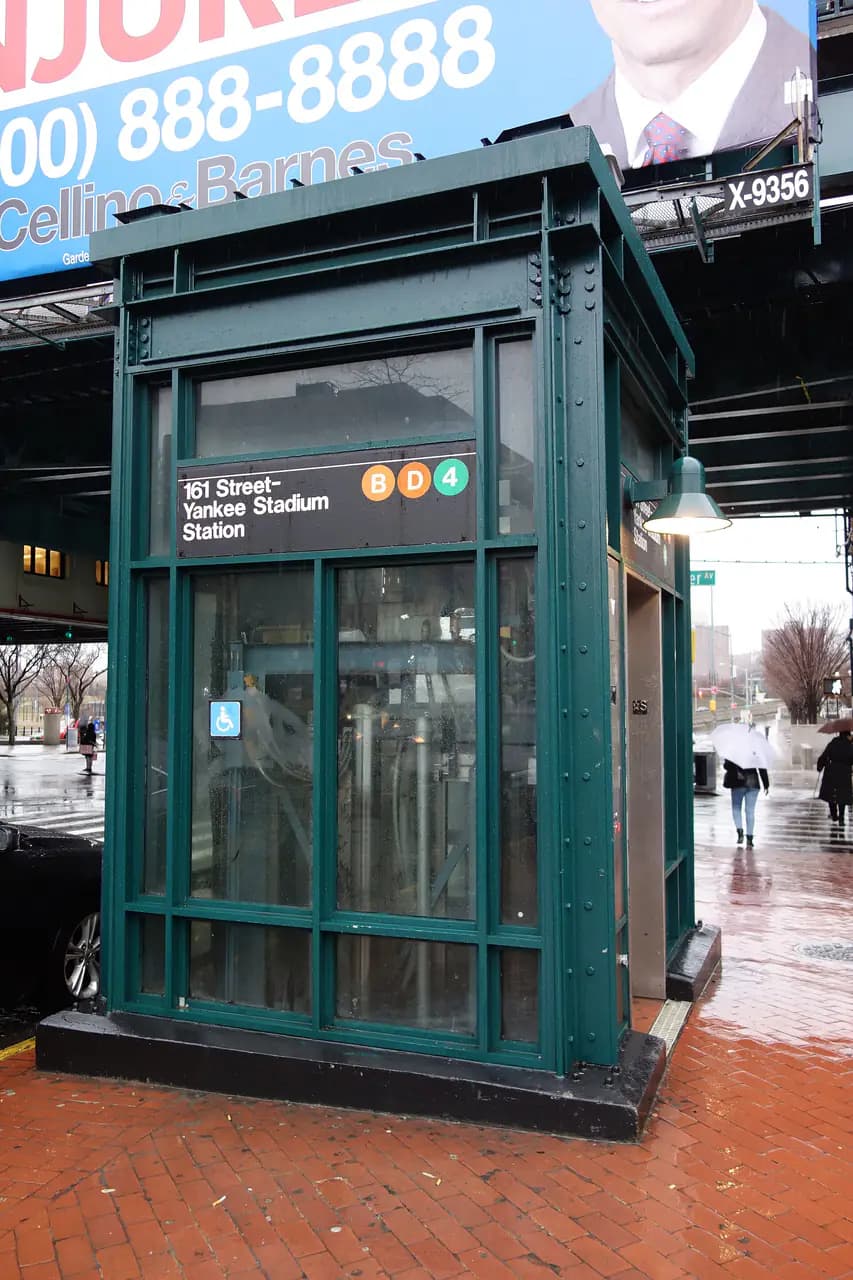 A green glass street elevator kiosk marked 161 Street-Yankee Stadium Station with B, D and 4 bullets, on a wet sidewalk under the elevated tracks