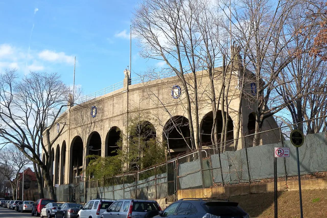 The arched concrete exterior of Forest Hills Stadium behind bare trees and a fence, with cars parked along a residential street