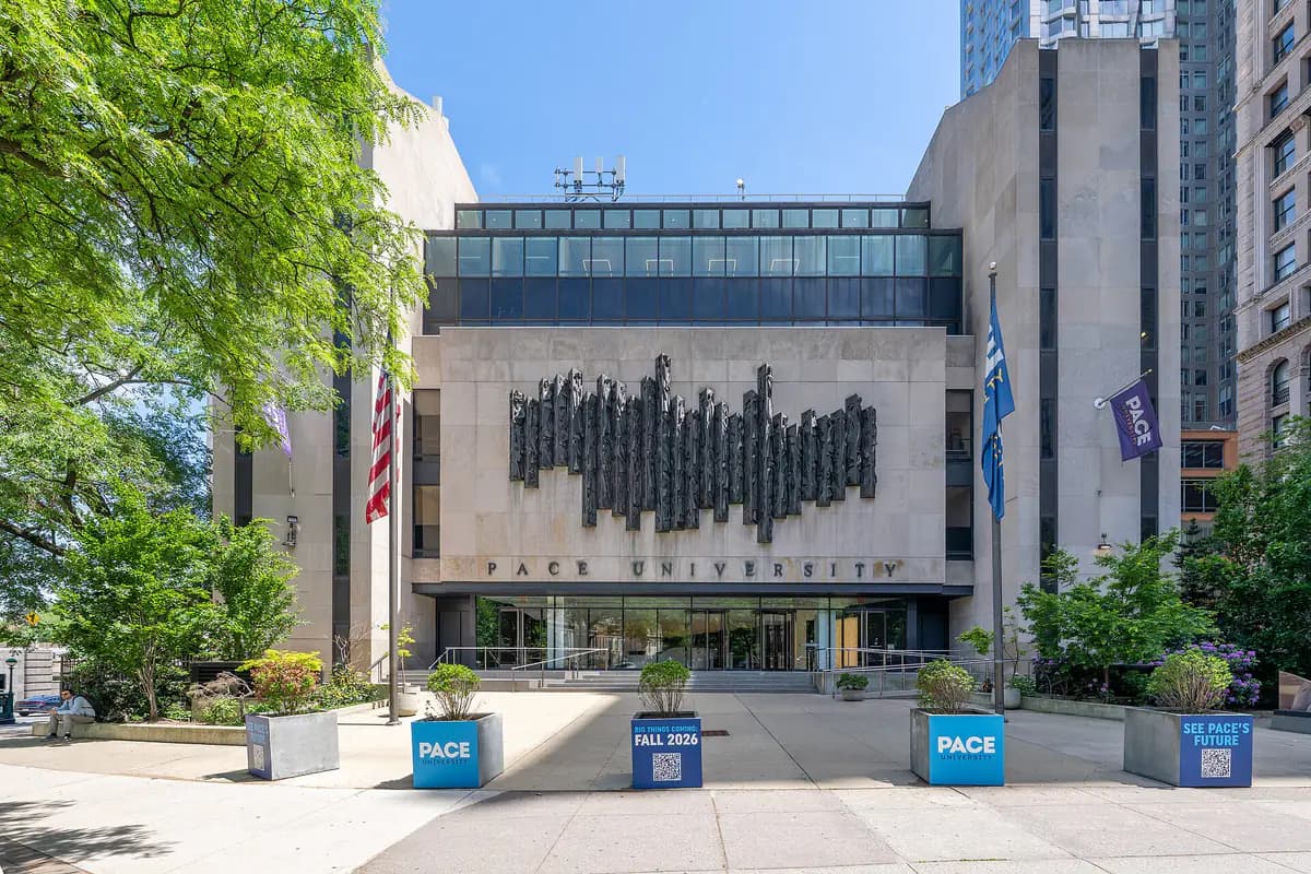The stone facade of One Pace Plaza with a dark metal sculpture over the entrance, flags and planters reading 'Pace' and 'Fall 2026'