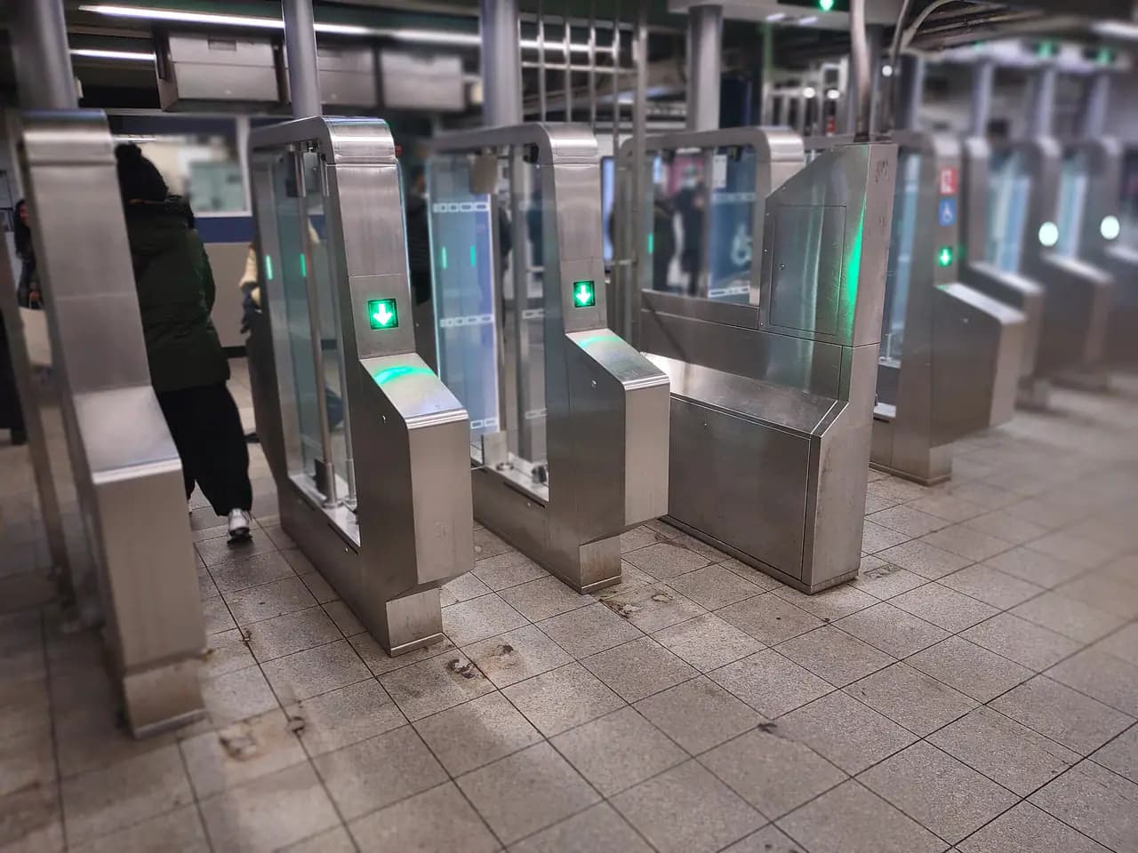 A row of new stainless-steel subway fare gates with glass doors and green arrow lights on a station mezzanine