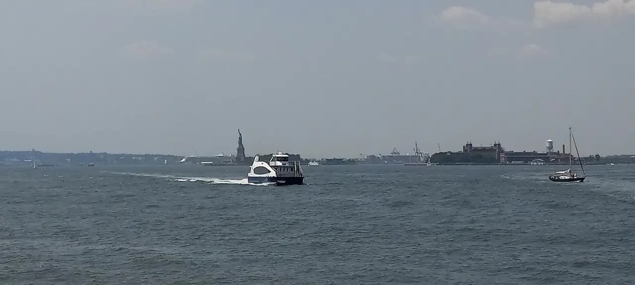 An NYC Ferry boat crossing New York Harbor off Battery Park City, with the Statue of Liberty and Ellis Island in the distance